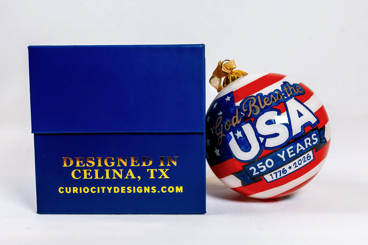God Bless the USA 250 Years hand-painted glass ornament beside its navy CurioCity Designs keepsake box.