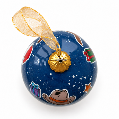 Overhead detail of A Celina Christmas glass ornament, showing its gold cap and ribbon, cowboy hat and wrapped gifts.