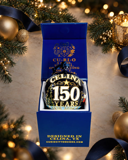 Celina 150 Years anniversary ornament in its CurioCity Designs gift box with navy ribbon, evergreen garland, and warm holiday lights.