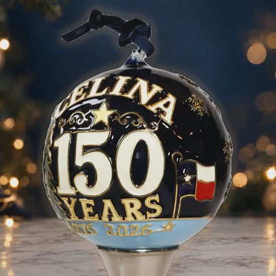 Glow-enhanced rotating Celina 150th Anniversary hand-painted glass ornament with anniversary lettering and longhorn artwork against a luxury Christmas background.
