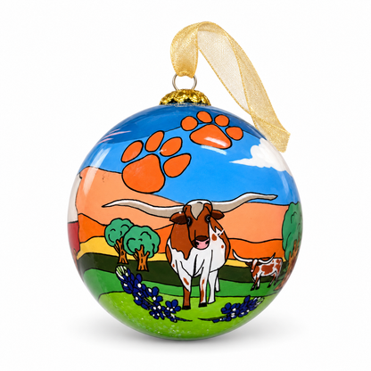 Celina Sunsets and Longhorn glass ornament styled with a gold ribbon, showing a longhorn, bluebonnets, orange paw prints, and a Texas sunset.