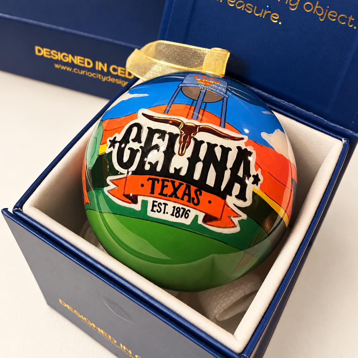 Reverse of the Celina Sunsets and Longhorn glass ornament with gold ribbon in its navy gift box, showing Celina, Texas and Est. 1876 lettering.